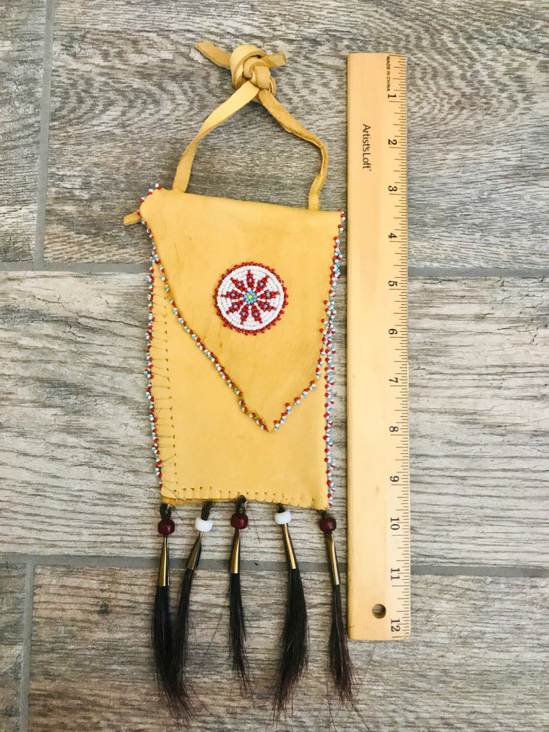 Puede incluir: Una peque&ntilde;a bolsa de cuero marr&oacute;n con un dise&ntilde;o de cuentas y flecos. La bolsa tiene un dise&ntilde;o de flor de cuentas en el centro y est&aacute; decorada con cuentas rojas, blancas y azules. La bolsa mide aproximadamente 18 cm de alto.