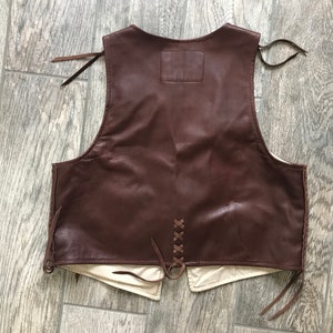May include: A brown leather vest with a lace-up back and fringe detailing. The vest has a plain back with no pockets or embellishments.