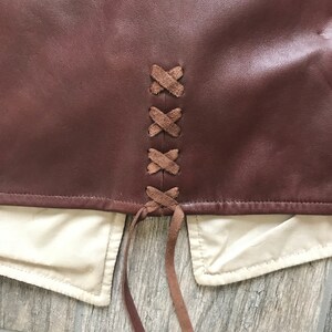 May include: A brown leather skirt with a lace-up back detail. The laces are brown and are tied in a knot at the bottom.