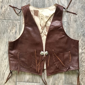 May include: A brown leather vest with fringe detailing, two pockets, and decorative stitching. The vest has a V-neck and ties at the front.