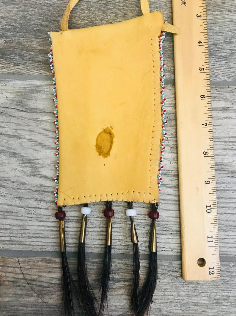 Puede incluir: Una peque&ntilde;a bolsa de cuero con cuentas y flecos de crin de caballo negro. La bolsa est&aacute; decorada con cuentas rojas, blancas y azules. Mide aproximadamente 15 cm de largo y 5 cm de ancho.