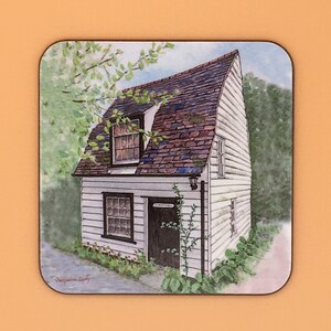 May include: A white cottage with a brown shingled roof and a black door. The cottage has a small window on the side and a larger window on the front. The cottage is surrounded by green trees and bushes.