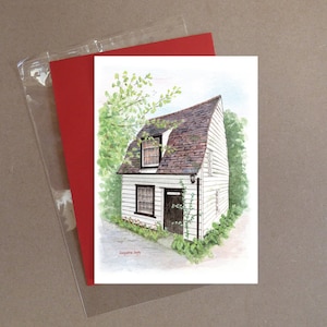 May include: A watercolour painting of a white cottage with a brown roof, black door and window frames, and green foliage. The artwork is on a white card with a red envelope in a clear plastic sleeve.