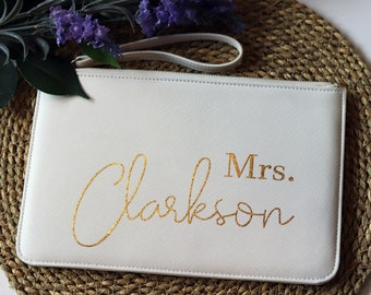 May include: A cream-coloured clutch bag with gold glitter lettering reading "Mrs. Clarkson". The bag has a wrist strap and is set on a woven mat, with greenery and purple flowers. A wedding accessory.