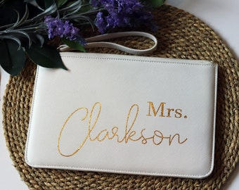 May include: A white clutch bag with a wrist strap, featuring the gold text "Mrs. Clarkson". The bag is on a woven mat with greenery and purple flowers in the background. A wedding accessory.