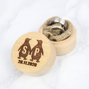 May include: A wooden ring box with a lid featuring two penguins with the initials "S" and "P" and the date "26.11.2020". The box is open and the lid is sitting on a white surface.