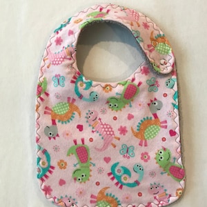 May include: Pink baby bib with a dinosaur and butterfly print. The bib has a gray fleece backing and a snap closure.