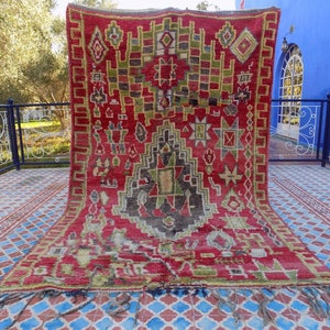 May include: A red, green, and black Moroccan rug with intricate geometric patterns and tassels. The rug is hanging on a wall with a blue background.