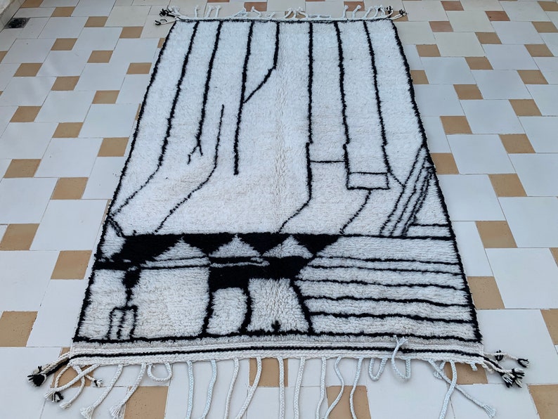 May include: A white and black Moroccan rug with a geometric pattern. The rug has a fringe border.