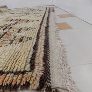 May include: A rectangular, cream-colored rug with a textured, woven design. The rug features brown and peach-colored geometric patterns and a fringed edge. The rug is placed on a white tiled floor.