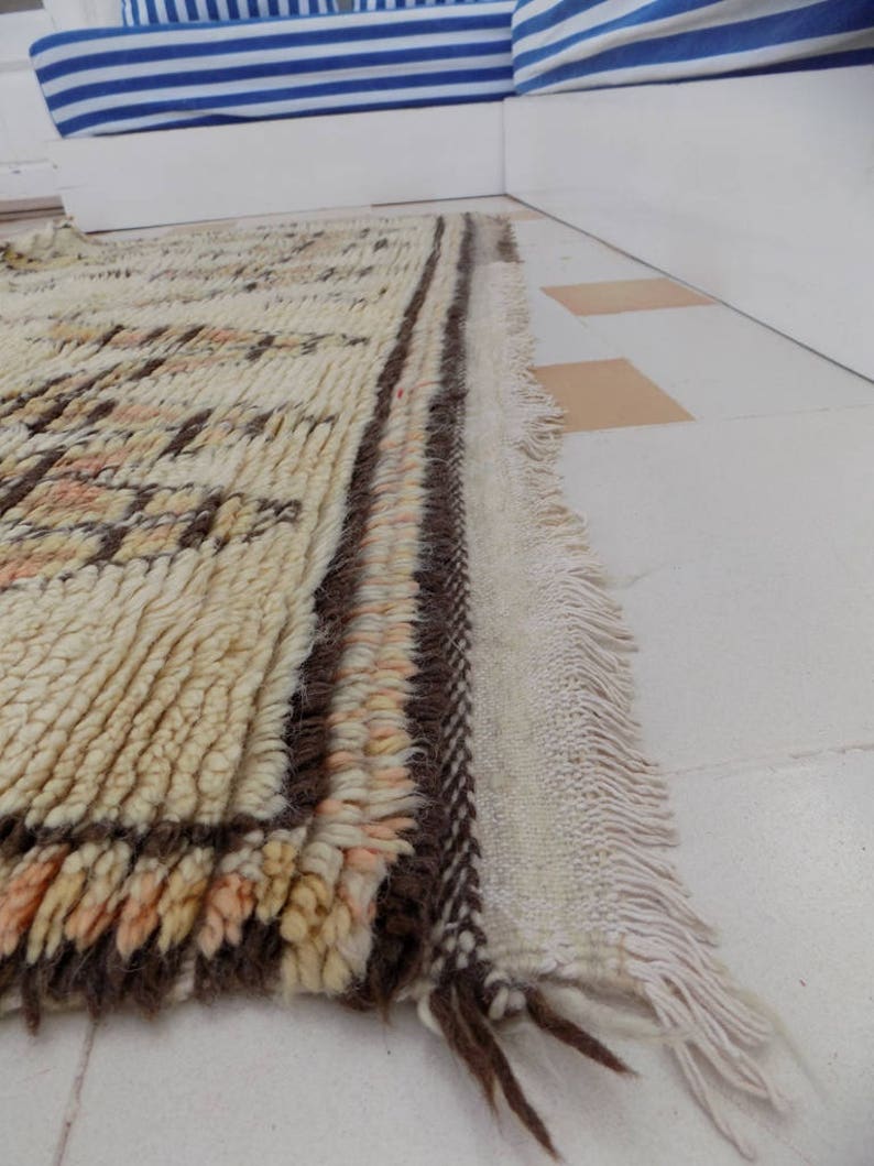 May include: A rectangular, cream-colored rug with a textured, woven design. The rug features brown and peach-colored geometric patterns and a fringed edge. The rug is placed on a white tiled floor.