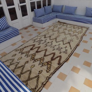 May include: A cream-colored, rectangular rug with a geometric pattern of brown zigzags and shapes. The rug is placed on a tiled floor, surrounded by white sofas with blue and white striped cushions. The scene is bright and airy.