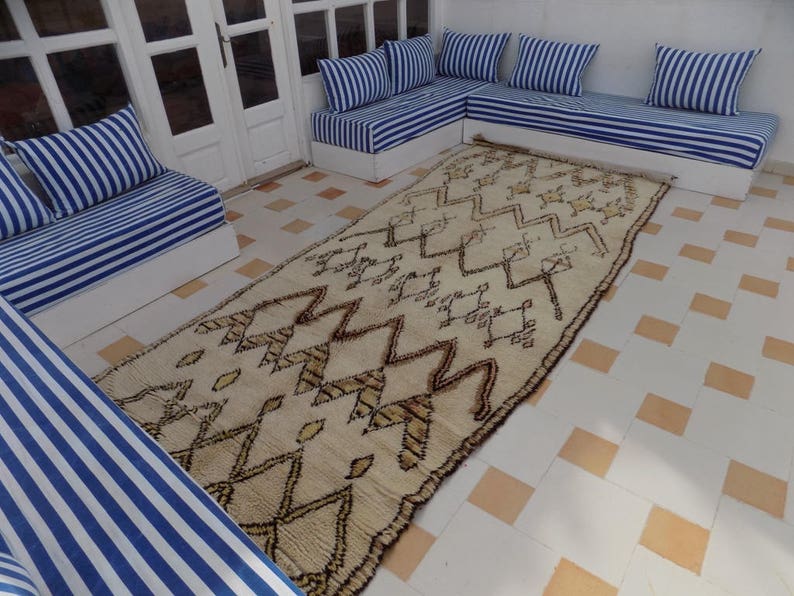 May include: A cream-colored, rectangular rug with a geometric pattern of brown zigzags and shapes. The rug is placed on a tiled floor, surrounded by white sofas with blue and white striped cushions. The scene is bright and airy.