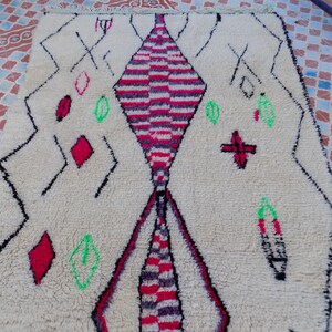 May include: A white Moroccan rug with a diamond pattern in the center. The diamond is made up of red, pink, and black stripes. The rug has a fringe border and is decorated with green and red geometric shapes.