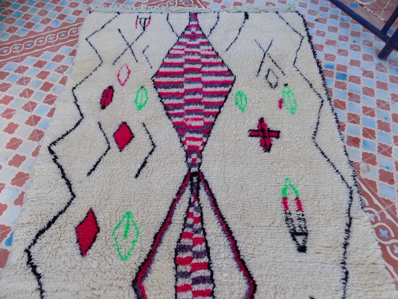 May include: A white Moroccan rug with a diamond pattern in the center. The diamond is made up of red, pink, and black stripes. The rug has a fringe border and is decorated with green and red geometric shapes.