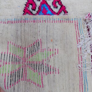 May include: Close-up of the back of a white rug with colorful woven details. The rug has a fringe border and is made of wool.