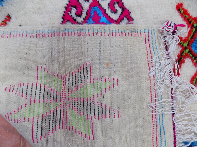 May include: Close-up of the back of a white rug with colorful woven details. The rug has a fringe border and is made of wool.