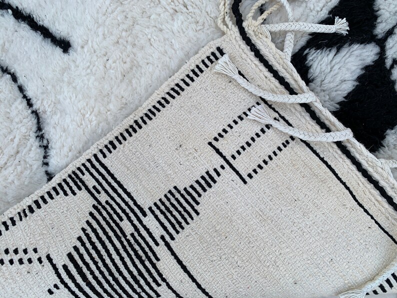 May include: A white and black geometric patterned rug with fringe. The rug has a simple design with black lines and squares on a white background.