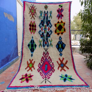 May include: A white Moroccan rug with a colorful geometric pattern. The rug has a fringe border and is hanging on a wall.