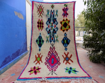 May include: A white Moroccan rug with a colourful geometric pattern. The rug has a fringe border and is hanging on a wall.