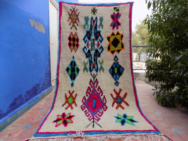 May include: A white Moroccan rug with a colorful geometric pattern. The rug has a fringe border and is hanging on a wall.