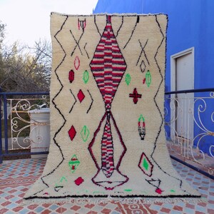May include: A white Moroccan rug with a large diamond pattern in red, black, and green. The rug has a fringe border and is hanging on a wall.