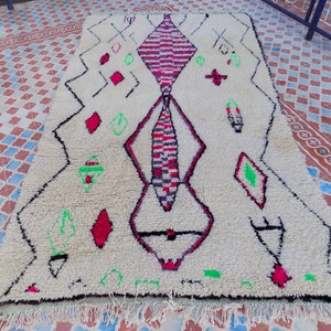 May include: A white Moroccan rug with a geometric design in black, red, and green. The rug has a fringe border.