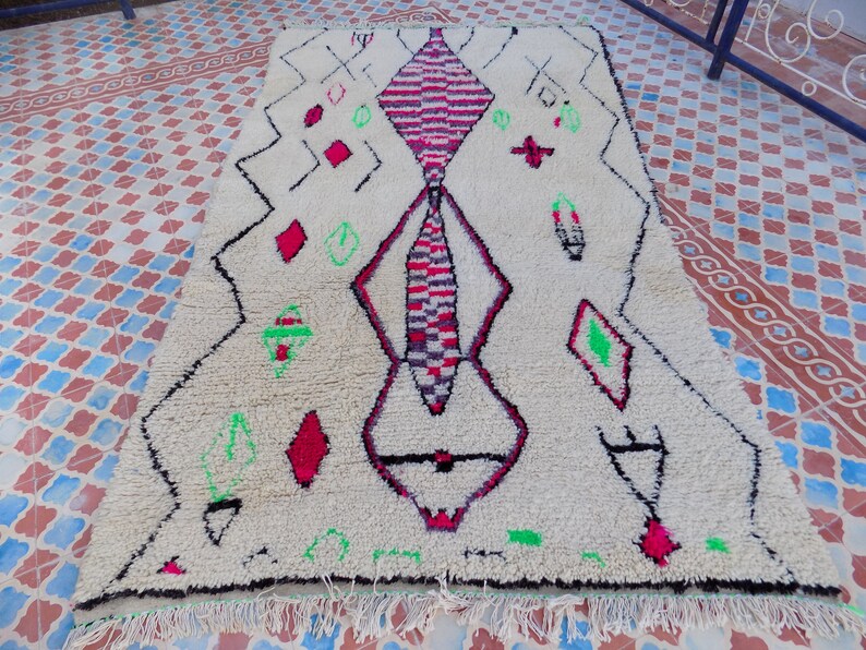 May include: A white Moroccan rug with a geometric design in black, red, and green. The rug has a fringe border.