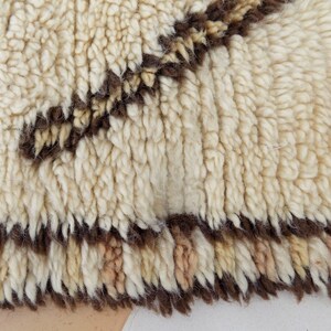 May include: Close-up of a cream-colored, textured rug with a brown geometric pattern. The rug has a thick, plush pile and a fringed edge. The design features a series of brown lines and shapes against the cream background.
