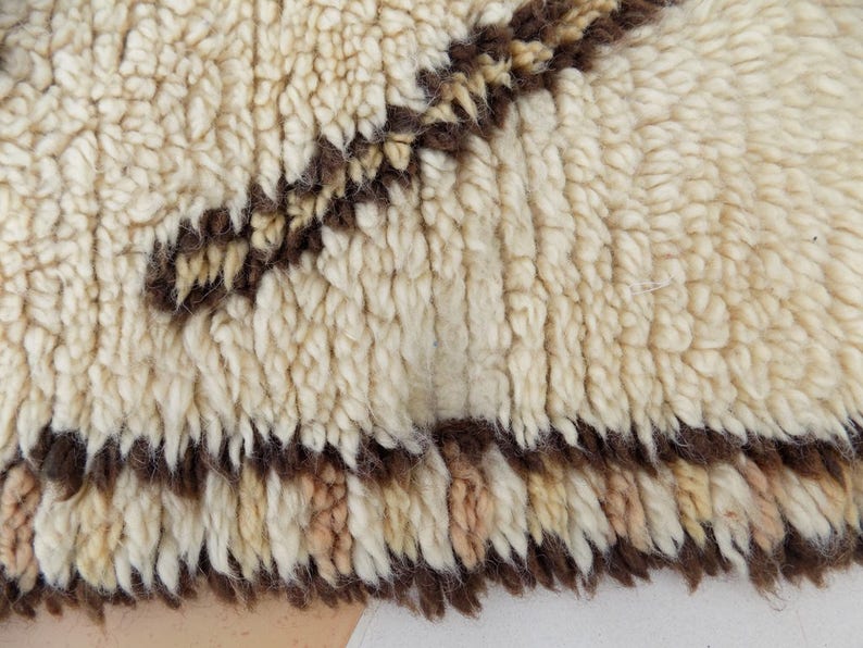 May include: Close-up of a cream-colored, textured rug with a brown geometric pattern. The rug has a thick, plush pile and a fringed edge. The design features a series of brown lines and shapes against the cream background.