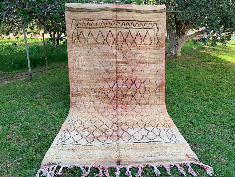 以下が含まれることがあります： 茶色、ピンク、クリームの色合いのダイヤモンド模様が施された、ビンテージのモロッコ製絨毯。フリンジの縁取りがあり、物干し竿にかけられています。