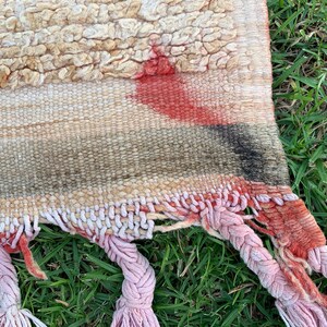 以下が含まれることがあります： ピンクと赤のフリンジが付いた織りの敷物のクローズアップ。敷物はクリーム色の背景に赤、黒、グレーの幾何学模様が施されています。