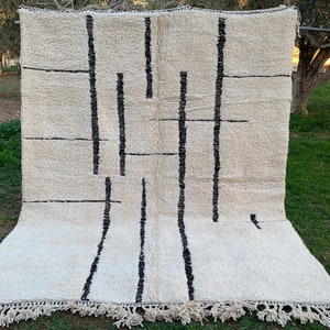 May include: A white rug with a black geometric pattern. The rug has a fringe border and is laid out on a grassy surface.