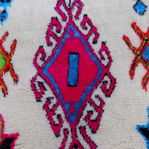 May include: A white rug with a colorful Moroccan design. The rug features a large diamond shape in the center with pink, blue, and black accents. The diamond shape is surrounded by smaller, intricate designs in white, pink, and blue. The rug also features smaller, star-shaped designs in red, green, and blue.