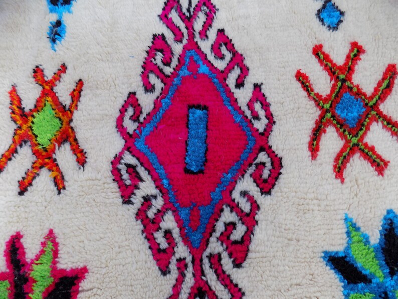 May include: A white rug with a colorful Moroccan design. The rug features a large diamond shape in the center with pink, blue, and black accents. The diamond shape is surrounded by smaller, intricate designs in white, pink, and blue. The rug also features smaller, star-shaped designs in red, green, and blue.