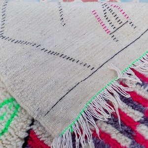 May include: Close-up of a white, cream, pink, and green Moroccan rug with a fringe border. The rug has a geometric pattern and is made of wool.