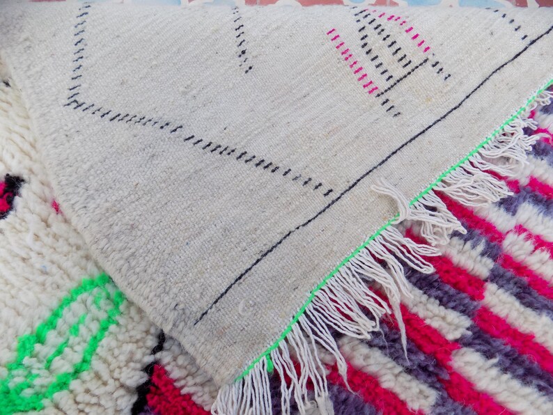 May include: Close-up of a white, cream, pink, and green Moroccan rug with a fringe border. The rug has a geometric pattern and is made of wool.