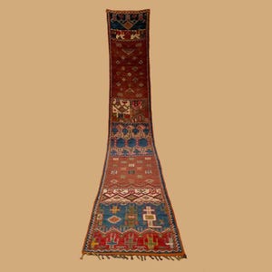 Puede incluir: Una alfombra larga y estrecha de forma rectangular con un patrón geométrico detallado. La alfombra presenta una paleta de colores rojo, azul y crema, con diseños intrincados y un borde con flecos. La alfombra es probablemente hecha a mano.