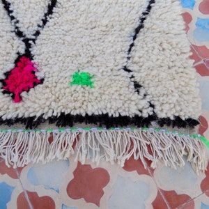 May include: A white wool rug with black, pink, and green geometric designs. The rug has a fringe border.