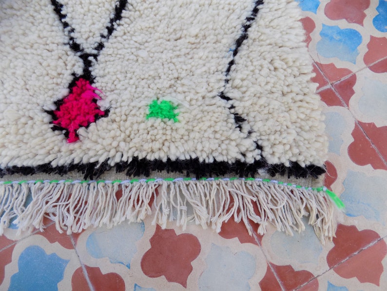 May include: A white wool rug with black, pink, and green geometric designs. The rug has a fringe border.