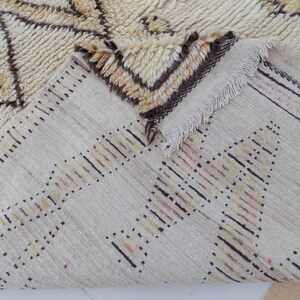 May include: Cream-colored rug with geometric patterns in brown and tan. The rug has a textured surface and a fringed edge. The design features a repeating pattern of triangles and lines, creating a cozy and inviting aesthetic.
