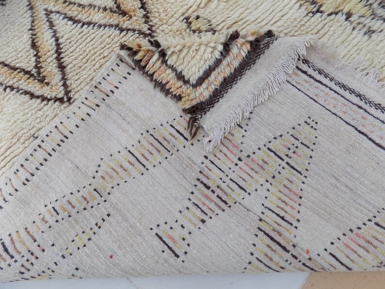 May include: Cream-colored rug with geometric patterns in brown and tan. The rug has a textured surface and a fringed edge. The design features a repeating pattern of triangles and lines, creating a cozy and inviting aesthetic.
