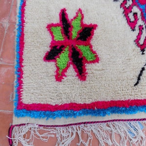 May include: White rug with a pink, green, and black floral design. The rug has a blue and pink border and white fringe.