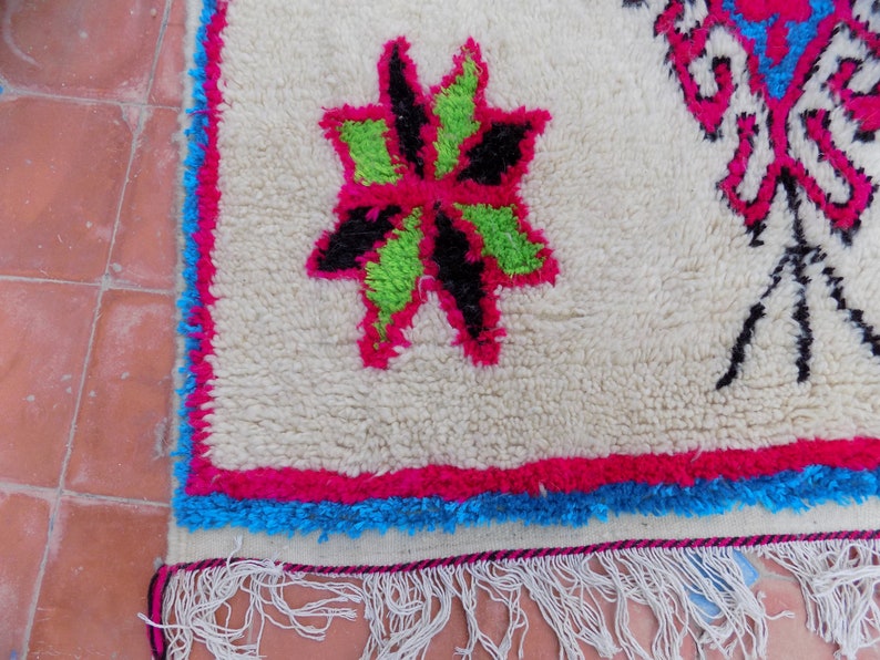 May include: White rug with a pink, green, and black floral design. The rug has a blue and pink border and white fringe.