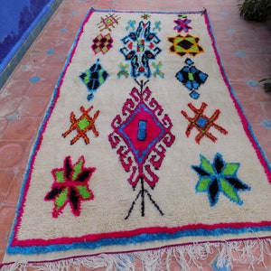May include: A white Moroccan rug with a colorful geometric pattern. The rug features a variety of shapes, including stars, diamonds, and squares. The colors are bright and vibrant, including pink, blue, green, and yellow.