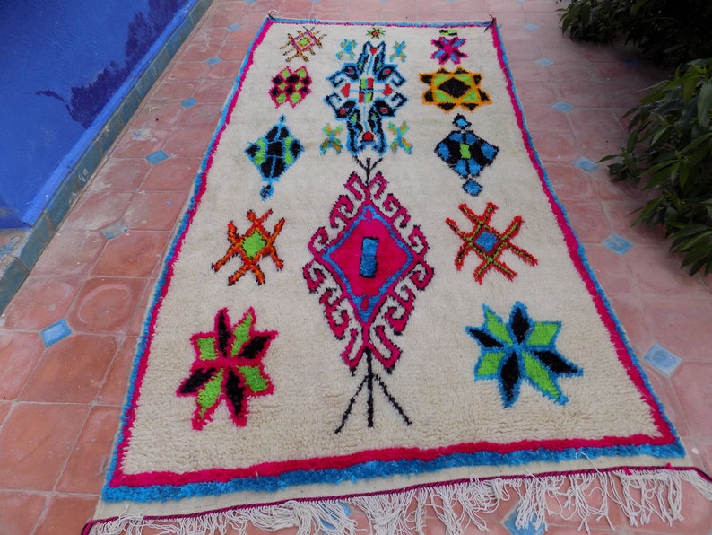 May include: A white Moroccan rug with a colorful geometric pattern. The rug features a variety of shapes, including stars, diamonds, and squares. The colors are bright and vibrant, including pink, blue, green, and yellow.