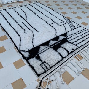 May include: White and black geometric patterned rug with fringe. The rug has a modern design and is made of wool.