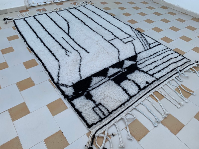 May include: White and black geometric patterned rug with fringe. The rug has a modern design and is made of wool.