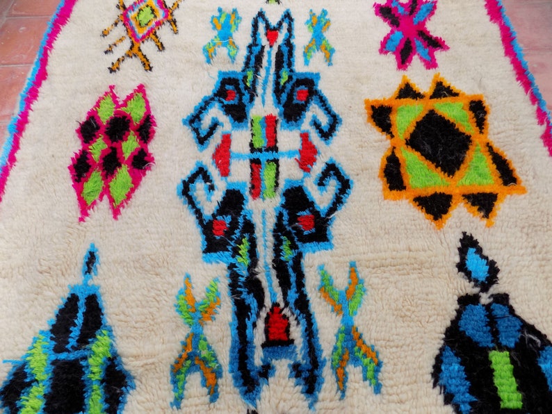 May include: A white rug with a colorful geometric pattern. The pattern features black, blue, green, red, orange, and yellow colors. The rug has a fluffy texture.