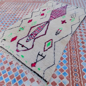 May include: A white Moroccan rug with a geometric pattern in shades of pink, red, green, and black. The rug has a fringe border and is laid out on a tiled floor.